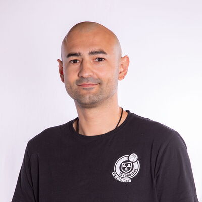 Ein Mann mit grauem Bart trägt ein T-Shirt mit bunten Logos und repräsentiert den Sportverein Turngemeinde Landshut.