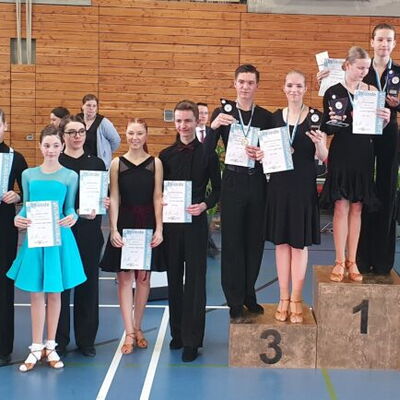 Tanzpaare der Turngemeinde Landshut posieren stolz mit Urkunden und Medaillen auf einem Siegerpodest in der Sporthalle.