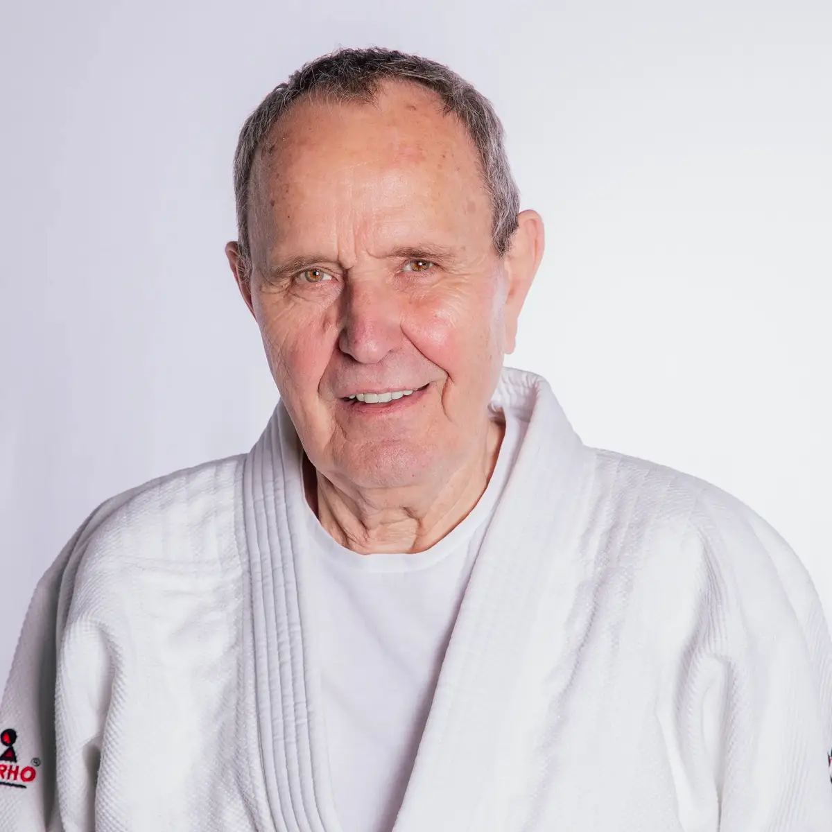 Ein älterer Mann in einem weißen Judogi lächelt freundlich in die Kamera, repräsentierend den Sportgeist der Turngemeinde.