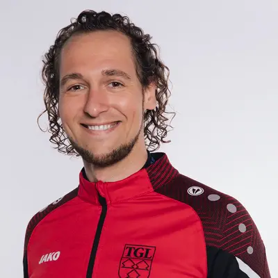 Ein Mann lächelt in einer roten TGL-Sportjacke, die das Vereinslogo der Turngemeinde Landshut zeigt.