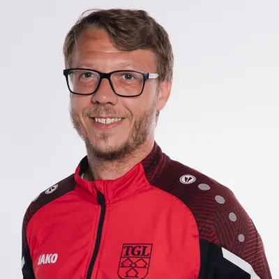 Ein Mann lächelt in einer roten TGL-Jacke, repräsentativ für den Sportverein Turngemeinde Landshut.