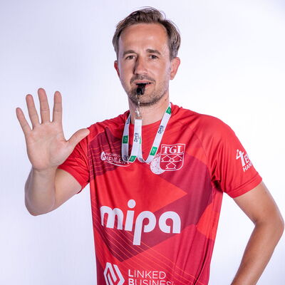 Ein Mann im roten TGL-Trikot hebt die Hand und trägt eine Pfeife, symbolisiert den Sportgeist des Vereins.