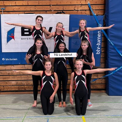 Eine Gruppe von Turnerinnen der TG Landshut posiert stolz in Sportkleidung mit einem Schild vor einem Turnhallenhintergrund.
