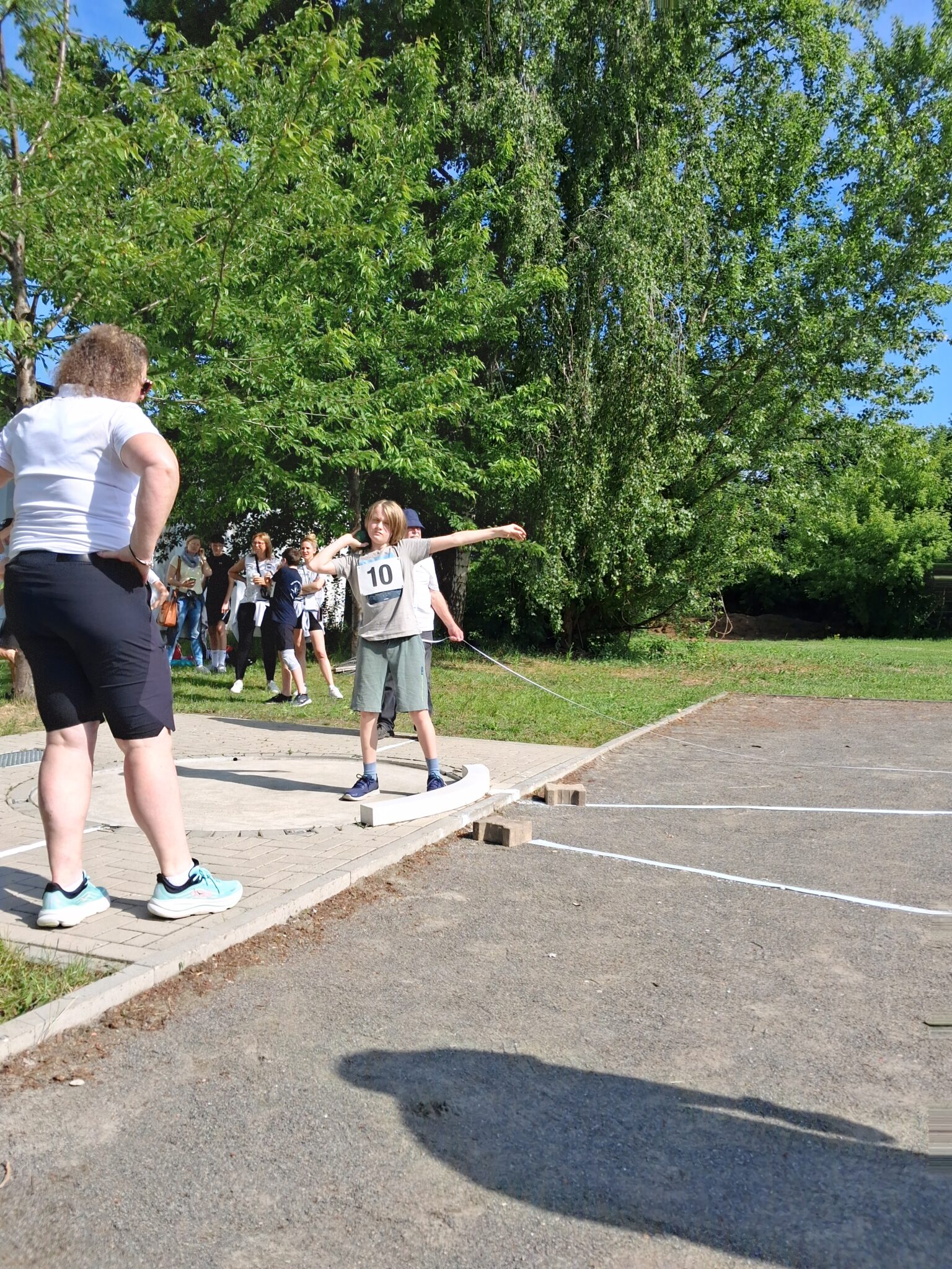 Ein Junge beim Kugelstoßen-Wettbewerb der Turngemeinde Landshut, beobachtet von einer Frau auf einem sonnigen Sportplatz.