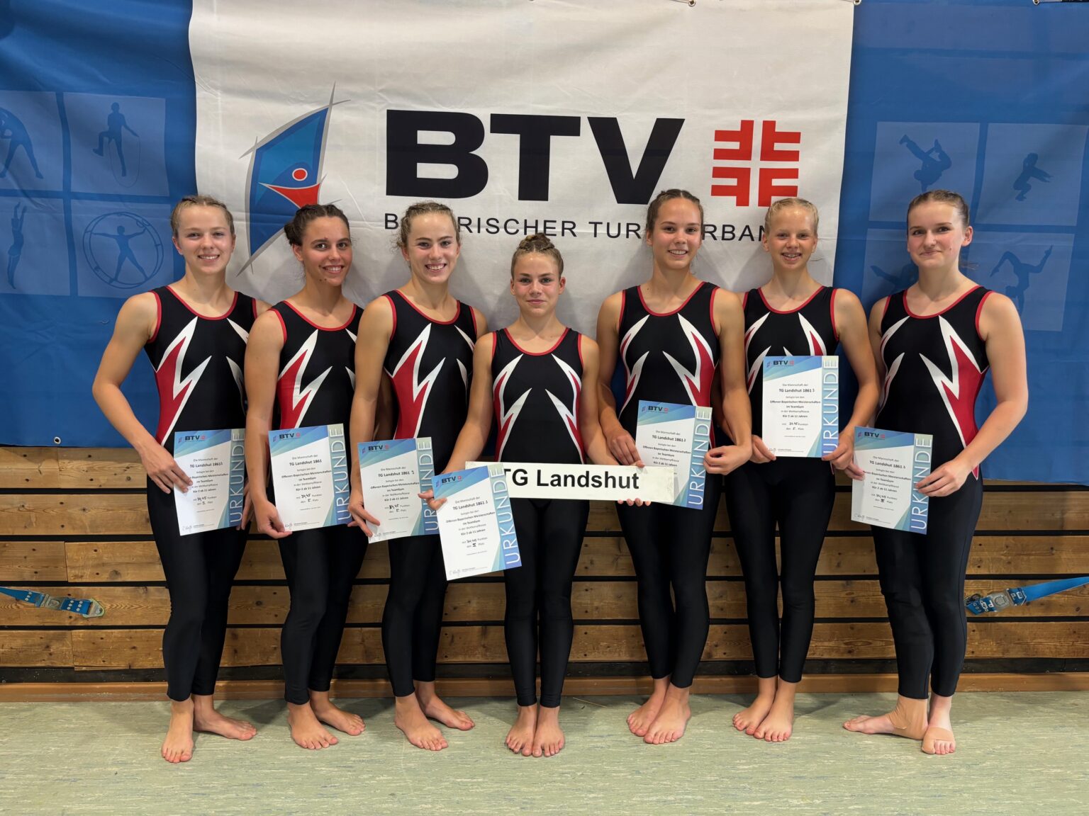Eine Gruppe von Turnerinnen der TG Landshut präsentiert stolz ihre Urkunden vor einem BTV-Banner.
