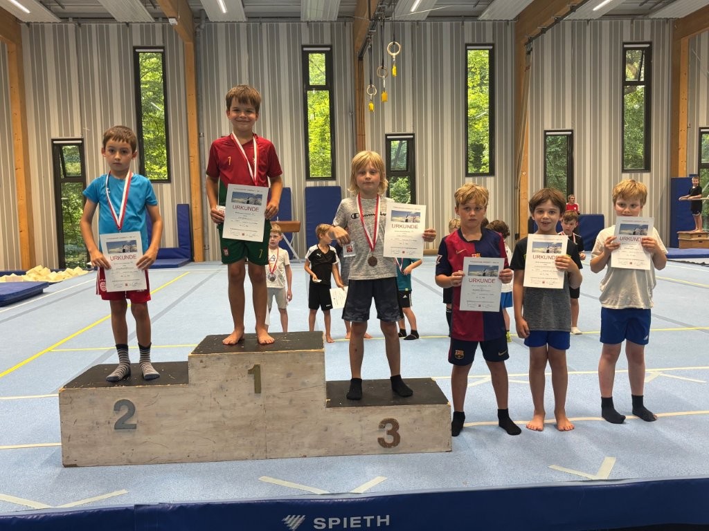 Sechs junge Jungs stehen auf einem Siegerpodest in einer Sporthalle des Vereins Turngemeinde Landshut und halten Urkunden.